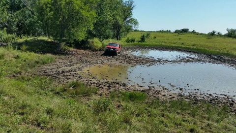 1986 Ford Bronco - A red SUV navigates a challenging muddy terrain with a pond nearby under a clear blue sky.
