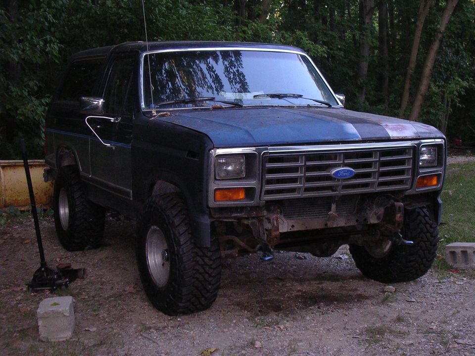 Lifted dark blue Ford Bronco on big mud tires with front bumper removed, parked on gravel near trees