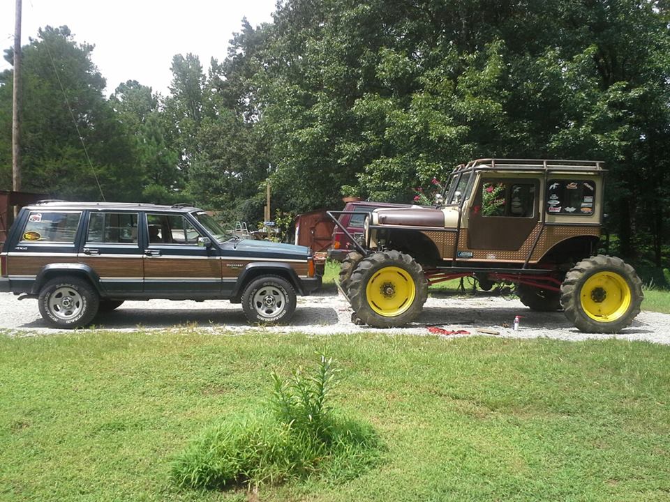 Custom lifted Jeep with large yellow tractor tires parked next to a wood-paneled SUV