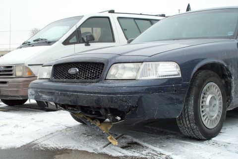 1998 Ford Crown Victoria - The front bumper of a Ford Crown Victoria is severely damaged and hanging loose, with a white van visible behind it.