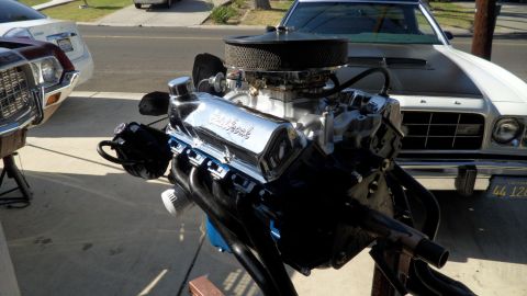 1972 Ford Ranchero - A chrome Edelbrock engine is displayed on a stand, with a white car a black stripe visible behind it.