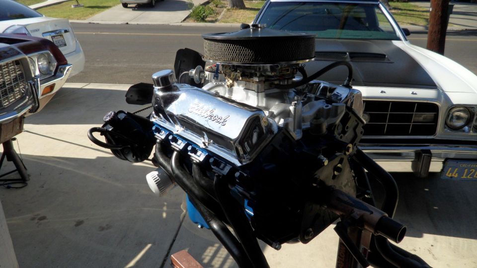 Chrome Edelbrock engine on stand with a white and black car in the background