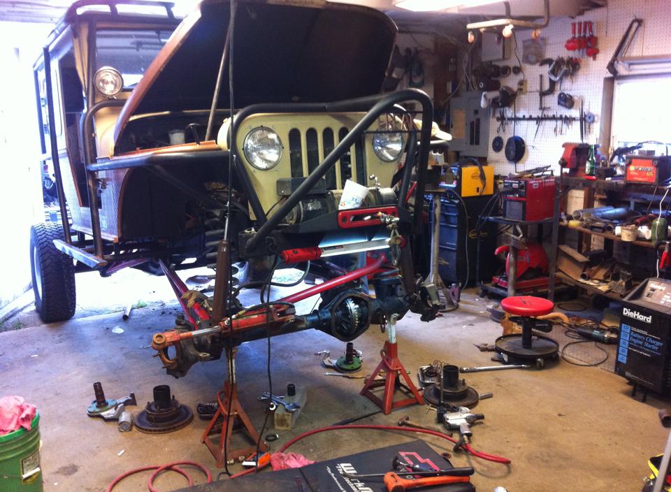 Jeep CJ-series in garage with hood up, front axle removed and supported by jack stands, tools and parts scattered around