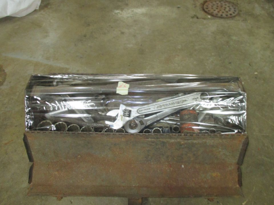 Metal tool organizer filled with wrenches and sockets, covered in plastic wrap