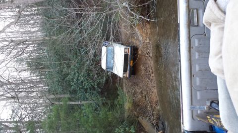 1979 Ford Bronco - A white SUV is stuck in a shallow creek, with trees and brush on either side of the bank.