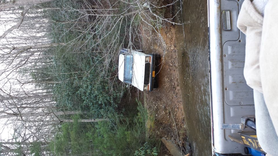 White SUV partially submerged in a creek bed, surrounded by trees