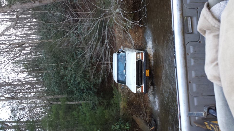 White 1978 Ford Bronco driving through a shallow stream in a wooded area