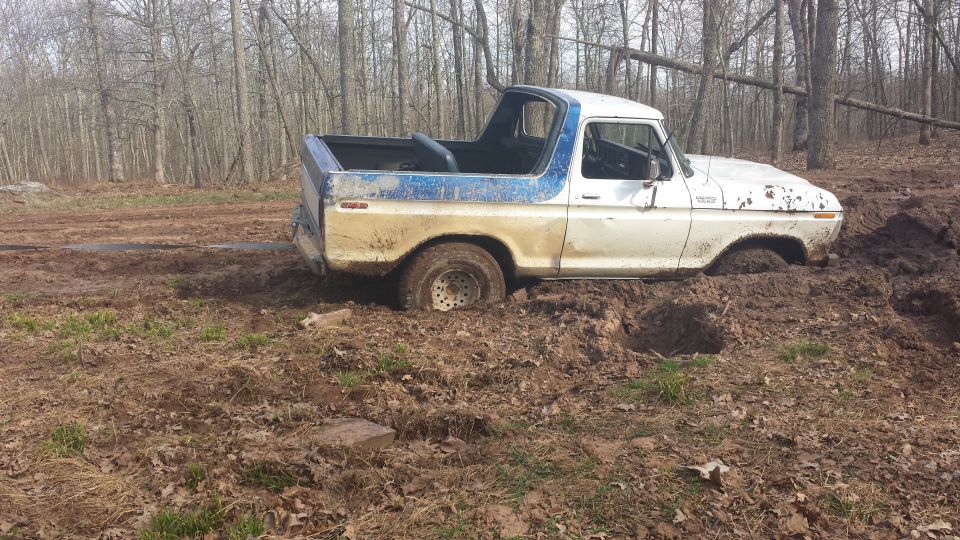 White and blue Ford Bronco stuck deep in mud in a wooded area
