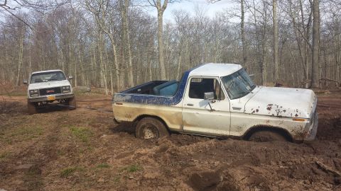 1979 Ford Bronco - Two trucks are stuck in the mud in a wooded area, with one attempting to tow the other.