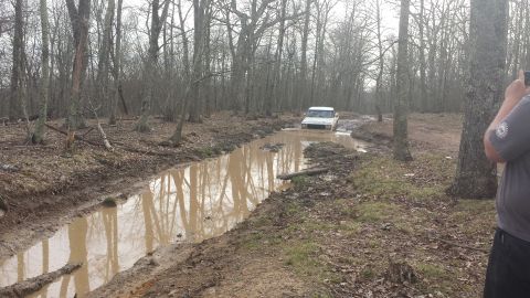 1979 Ford Bronco - A white SUV navigates a deep mud-filled trench in a wooded area, with a person filming from the side.