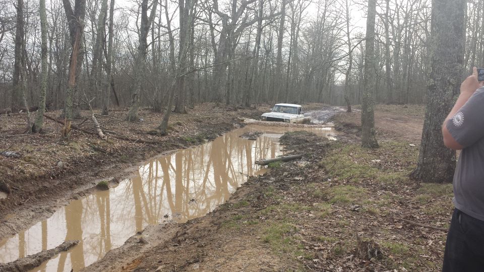 White 1978 Ford Bronco driving through a muddy ditch in a forest