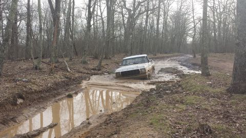 1979 Ford Bronco - A white Ford pickup truck navigates a deep mud puddle on a wooded trail.