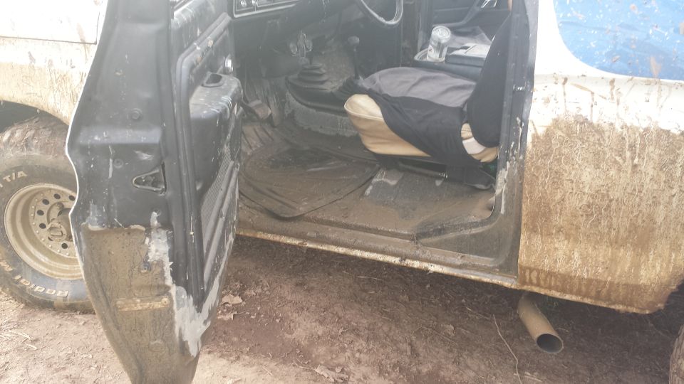 Mud-splattered 1978 Ford Bronco interior with open driver's door, showing floor mats, seats, and gear shifter