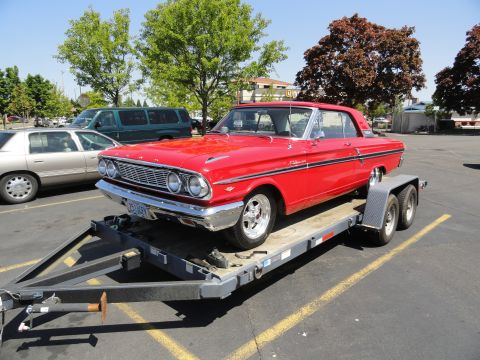 1964 Ford Fairlane - A bright red 1964 Ford Fairlane is being transported on a trailer, parked in a lot.