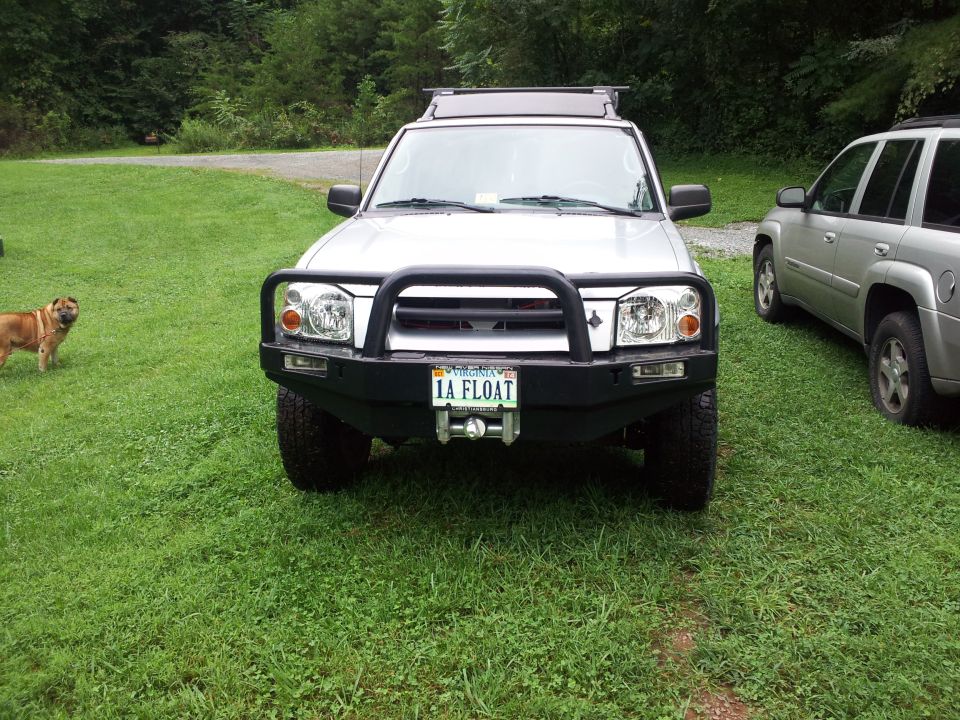 Front view of a silver Nissan Frontier pickup with a custom bumper and bull bar. A dog stands in the grass to the left, and another SUV is parked to the right