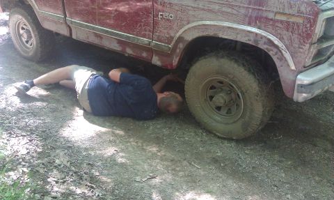 1979 Ford Bronco - A person works on the underside of a muddy red pickup truck, possibly a 1979 Ford F-150.
