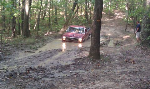 1979 Ford Bronco - Red pickup truck stuck in mud on a wooded trail. The grille suggests a 1980s model.