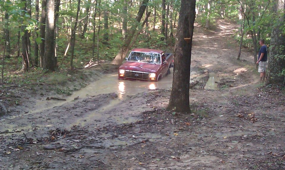 Red pickup truck stuck in mud on a wooded trail. The grille suggests a 1980s model