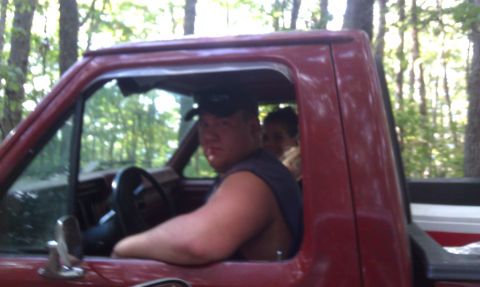 1979 Ford Bronco - Two people are inside a red pickup truck, parked in a wooded area.