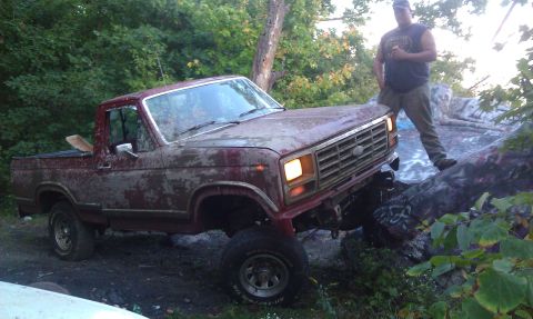 1979 Ford Bronco - A man stands next to a weathered red pickup truck as it navigates a challenging rocky terrain.