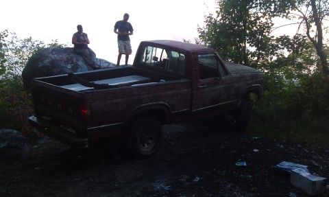 1979 Ford Bronco - A weathered red pickup truck sits off-road with two men observing.