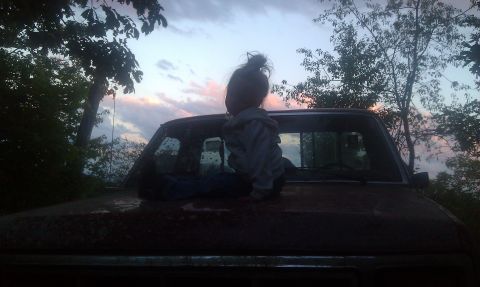 1979 Ford Bronco - A child sits on the hood of a pickup truck as the sun sets, with trees and clouds in the background.
