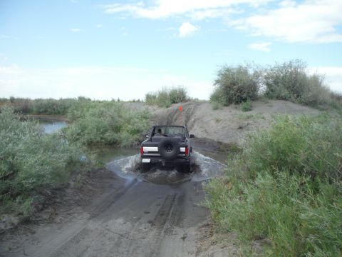 2005 Jeep Wrangler - A black convertible SUV navigates through a shallow body of water on a dirt trail surrounded by vegetation.