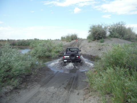 2005 Jeep Wrangler - Jeep Wrangler navigates a water obstacle on a dirt path, with vegetation on either side.