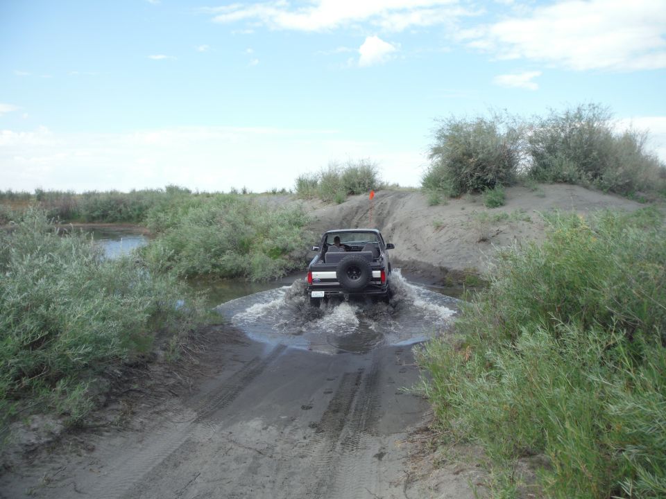 2005 Jeep Wrangler driving through a shallow water crossing on a dirt track, surrounded by brush