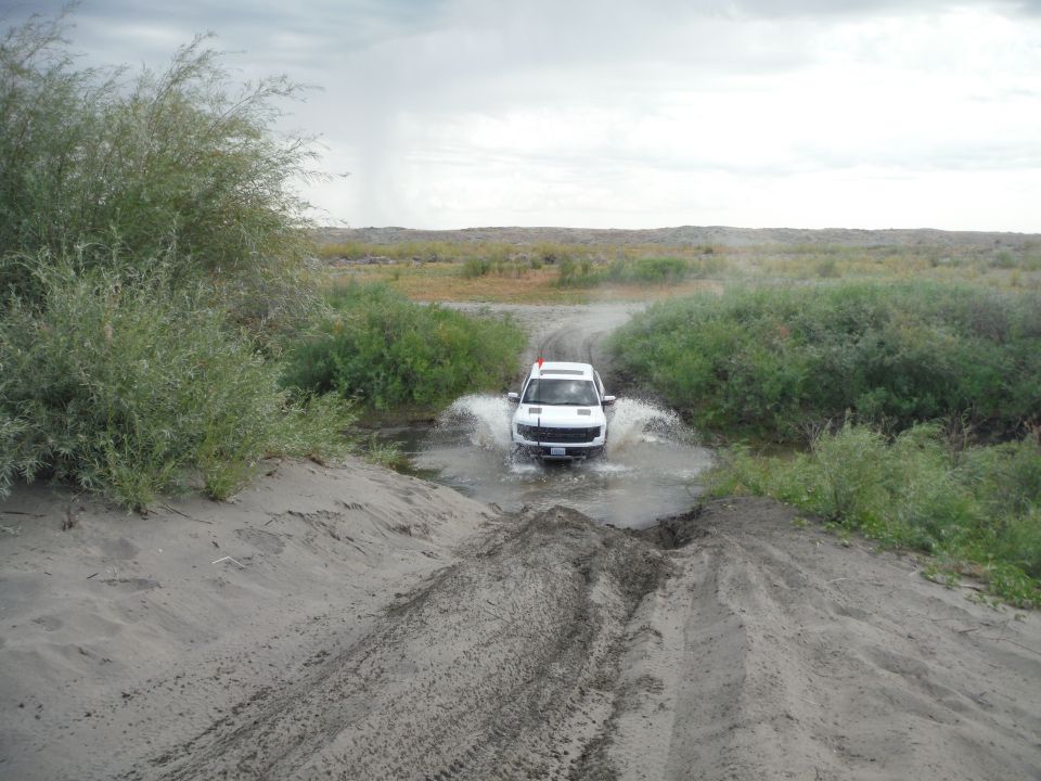 White SUV splashing through a shallow stream on a dirt path, with sand dunes and brush on either side