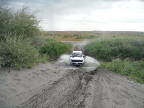 2005 Jeep Wrangler - A white SUV navigates a water hazard on a dirt track, kicking up spray.