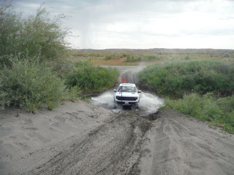 2005 Jeep Wrangler - A white SUV splashes through a muddy puddle on a dirt road, surrounded by dry vegetation and a cloudy sky.