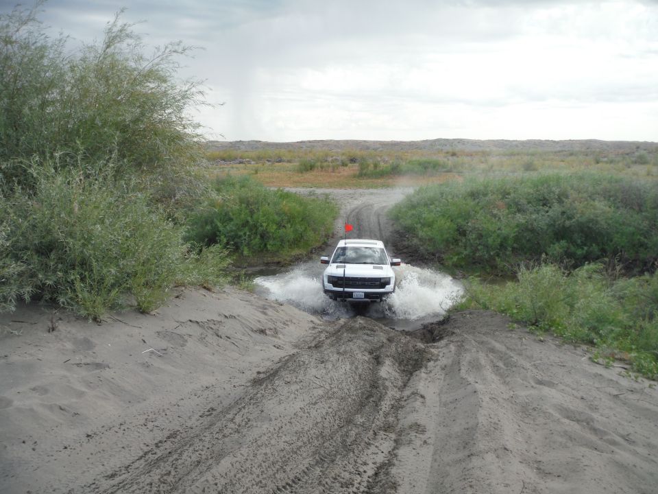 White pickup truck splashing through a muddy puddle on a dirt track, with brush and dry landscape in the background