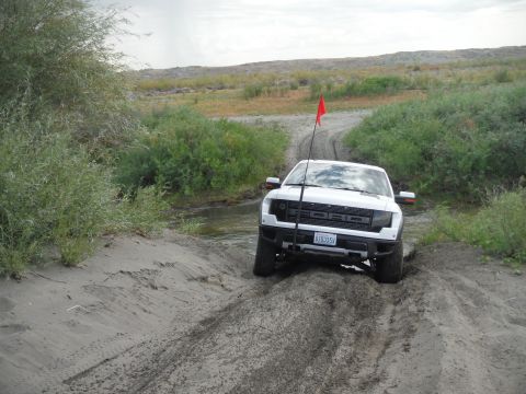 2005 Jeep Wrangler - White Ford Raptor truck navigates a sandy, off-road path with vegetation and a small stream nearby.