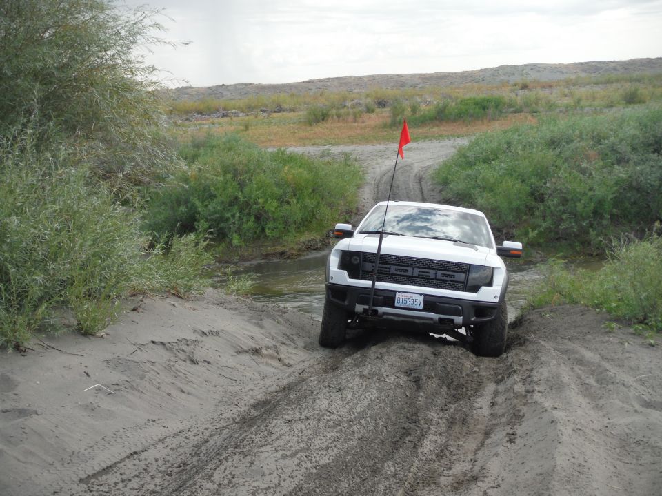 White Ford Raptor truck with a red flag on a sandy dirt road near bushes and a stream