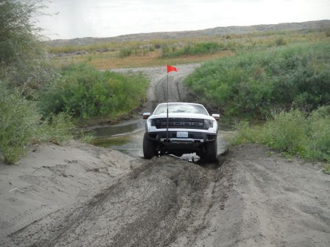 2005 Jeep Wrangler - A white Ford Raptor truck navigates a muddy off-road path, with vegetation on either side and a red flag mounted on the front.