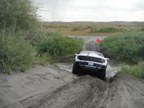 2005 Jeep Wrangler - A white Ford F-150 Raptor navigates a challenging muddy and watery off-road path under an overcast sky.