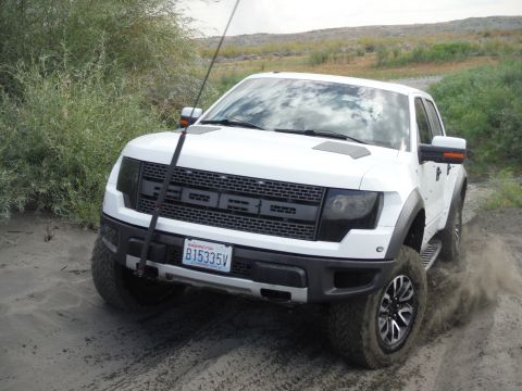 2005 Jeep Wrangler - A white Ford F-150 Raptor kicks up dust as it drives on a dirt road, its distinctive grille clearly visible.