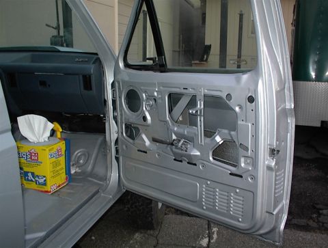 1990 Ford Bronco - The interior of a silver vehicle is shown with the driver's door open, revealing the removed door panel and a box of rags on the floor.