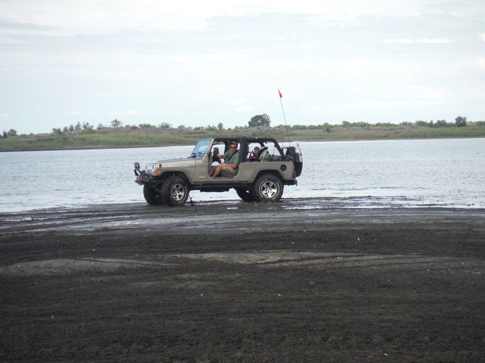 Tan 2005 Jeep Wrangler with people inside parked on a dark sandy area near a body of water