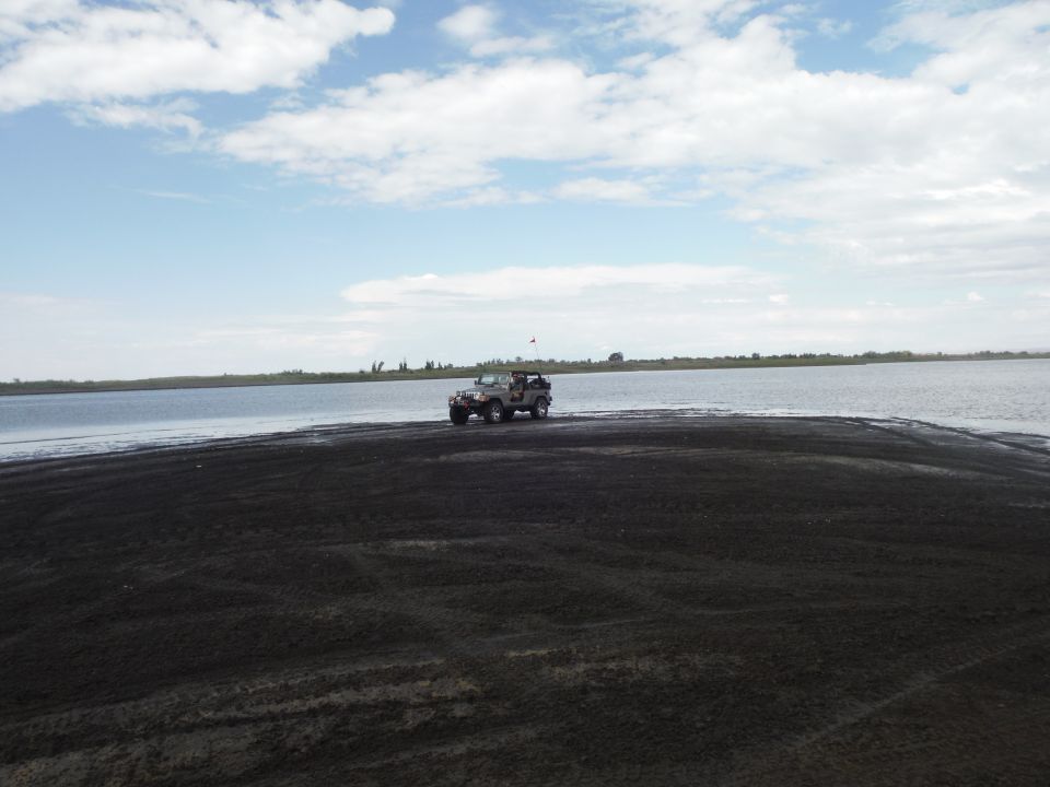 Gray Jeep Wrangler on dark sand near water, with a flag on the antenna