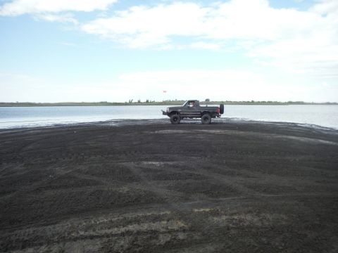 2005 Jeep Wrangler - Ford Bronco sits on a dark sandy shore with a calm lake in the background under a cloudy sky.