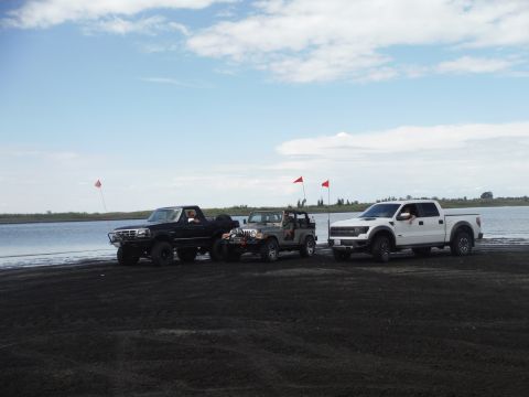 2005 Jeep Wrangler - A black pickup, a gray Jeep, and a white pickup truck are parked on a dark sandy shore near a lake under a cloudy sky.
