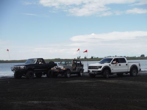 2005 Jeep Wrangler - Three off-road vehicles are parked on the dark sand near a lake under a cloudy sky.