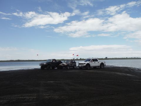 2005 Jeep Wrangler - Multiple off-road vehicles are parked on a dark, sandy area next to a calm body of water.