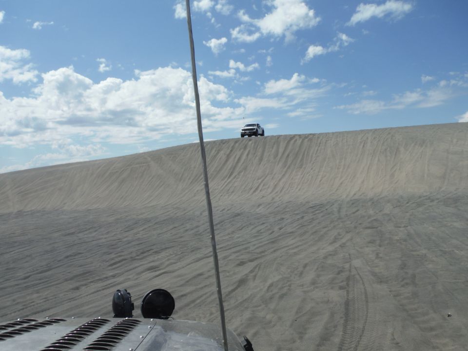 White SUV on a sand dune under a blue sky with clouds. Another vehicle's hood is in the foreground
