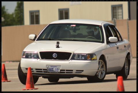 2000 Mercury Grand Marquis - A white 2000 Mercury Grand Marquis sedan navigates a course marked by orange traffic cones, with a driver wearing a helmet visible inside.