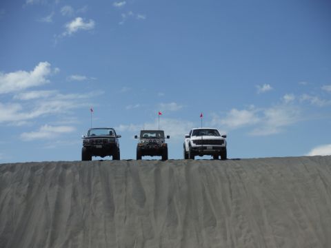 2005 Jeep Wrangler - Three off-road vehicles are positioned on a sandy ridge against a bright blue sky with scattered clouds.