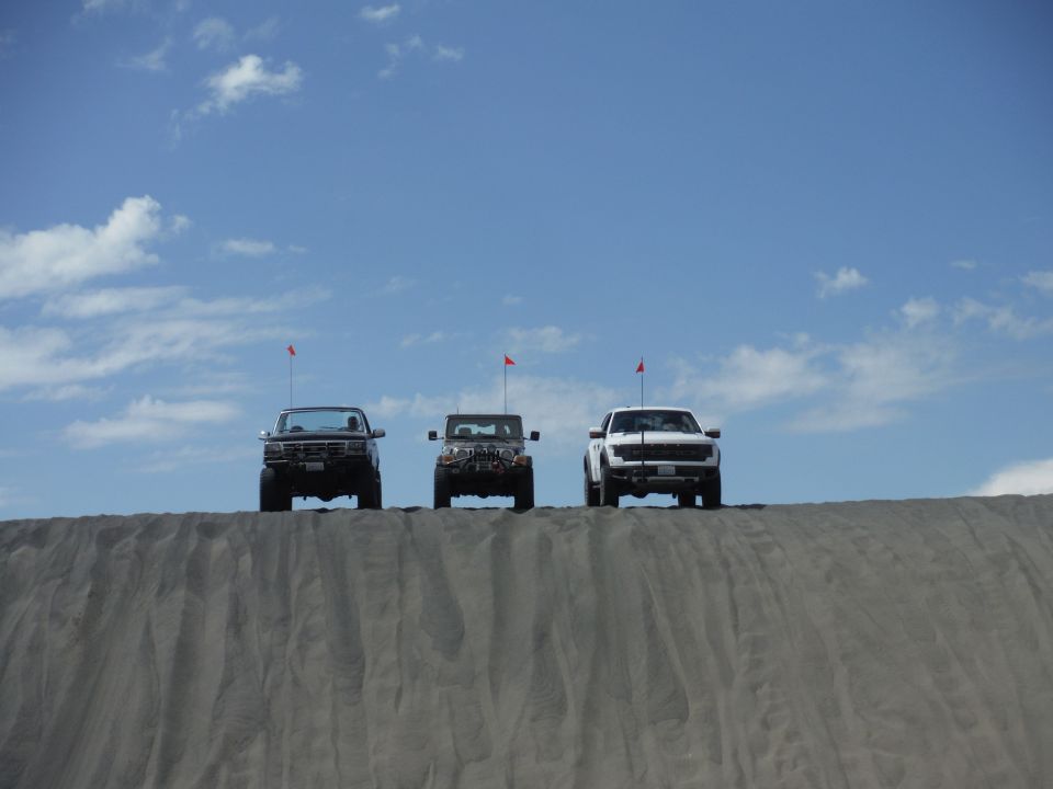 Three trucks and SUVs are lined up on top of a sand dune under a blue sky with some clouds. Each vehicle has a red flag on an antenna