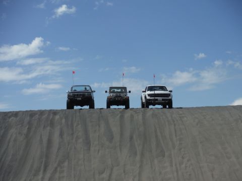 2005 Jeep Wrangler - A white Ford F-150 Raptor, a dark Jeep Wrangler, and a dark SUV are positioned on a sand dune against a backdrop of clouds.